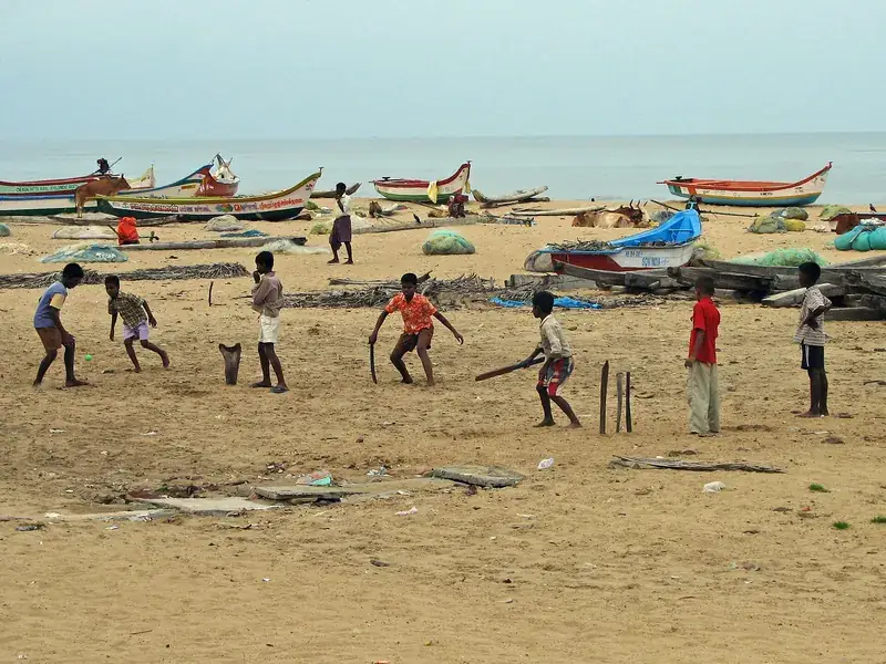 Children playing cricket on a beach beside fishing boats.
